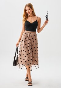 Schwarzes Top mit Muschelsaum, kombiniert mit einem transparenten beigen Rock mit schwarzen Herzmustern. Das Modell trägt eine schwarze Handtasche und eine Sonnenbrille.