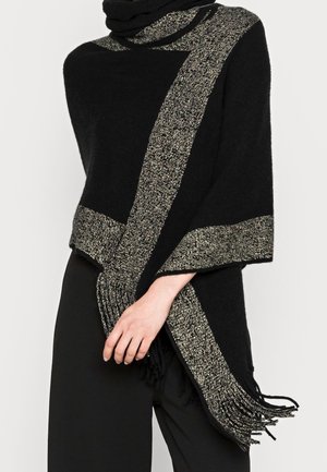Poncho noir avec de larges rayures métalliques argentées tricotées le long des bords et des extrémités à franges, porté sur un pantalon noir.