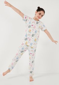Puuvillasta valmistettu pyjama-setti, joka sisältää lyhythihaisen paidan ja elastiset housut. Monivärisiä sarjakuvahahmoja ja sateenkaaria valkoisella taustalla.