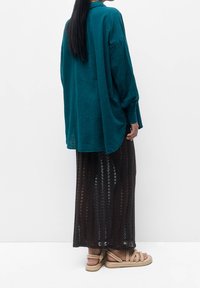 Chemise à manches longues bleu sarcelle avec un col, associée à une jupe plissée noire transparente et des sandales tressées. Tissu léger et texturé.