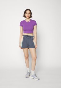 Violet trumpas marškinėliai, derinami su tamsiai pilkomis šortais, turinčiais pastelinių spalvų panelius. Modelis dėvi baltas kojines ir šviesiai pilkus sportinius batus.