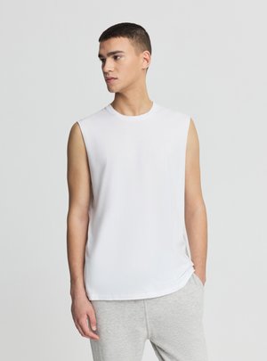 Giovane uomo in piedi con le mani nelle tasche dei pantaloni grigi, indossa una canottiera bianca, guarda verso sinistra su uno sfondo semplice.