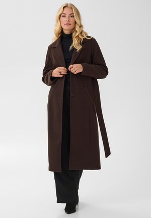 Femme aux longs cheveux blonds portant un manteau long marron foncé sur un col roulé noir et un pantalon, debout devant un fond gris uni.