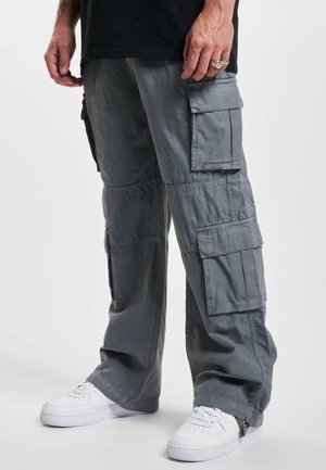Homme portant un pantalon cargo gris ample avec plusieurs poches et des baskets blanches, debout devant un fond clair uni.