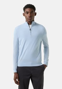 Lichtblauwe trui met rits, gemaakt van een zachte, gestructureerde brei. Voorzien van een hoge kraag en geribbelde manchetten. Gecombineerd met een donkere broek.