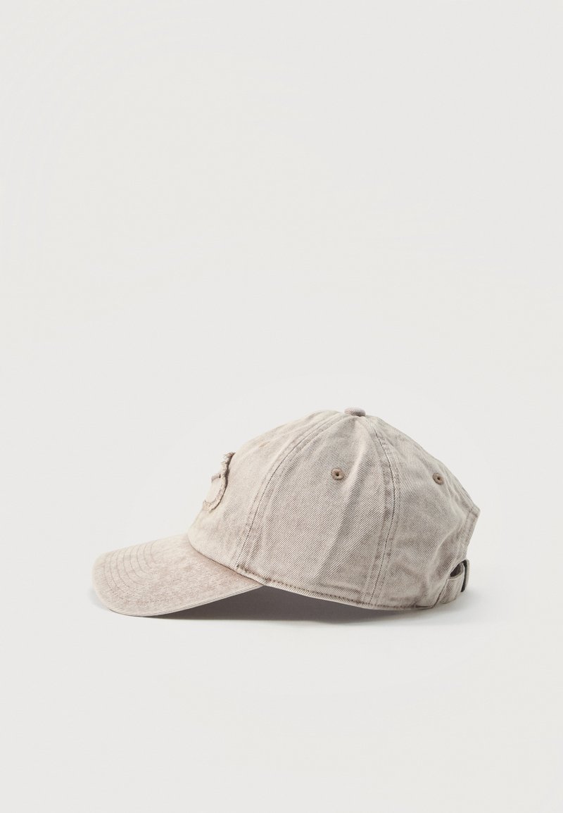 Gorra de béisbol de algodón beige con paneles cosidos, ojales de ventilación y visera curvada, mostrada desde el lado izquierdo sobre un fondo liso.