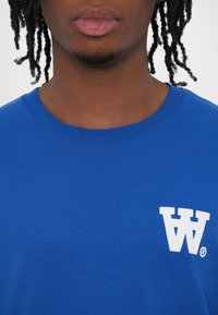 Blue cotton t-shirt featuring a ribbed crew neckline, short sleeves, and a white logo on the left chest area. Smooth texture.