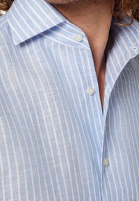 Jasnoniebieska koszula z białymi pionowymi paskami, zapinanym kołnierzykiem i częściowo rozpiętym przodem, na osobie z kręconymi włosami.