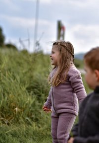Lyse lilla fleeceoutfit med lynlås og manschetter ved anklerne, båret af et barn med langt, bølget hår og en fletning, udenfor i græsbevoksede omgivelser.
