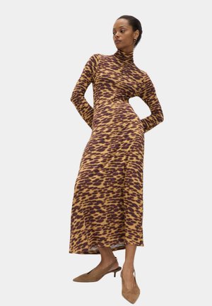 Langes, figurbetontes Kleid in Braun und Gelb mit Animal-Print. Hoher Hals, lange Ärmel, weicher Stoff. Modell posiert selbstbewusst mit Händen in den Taschen.