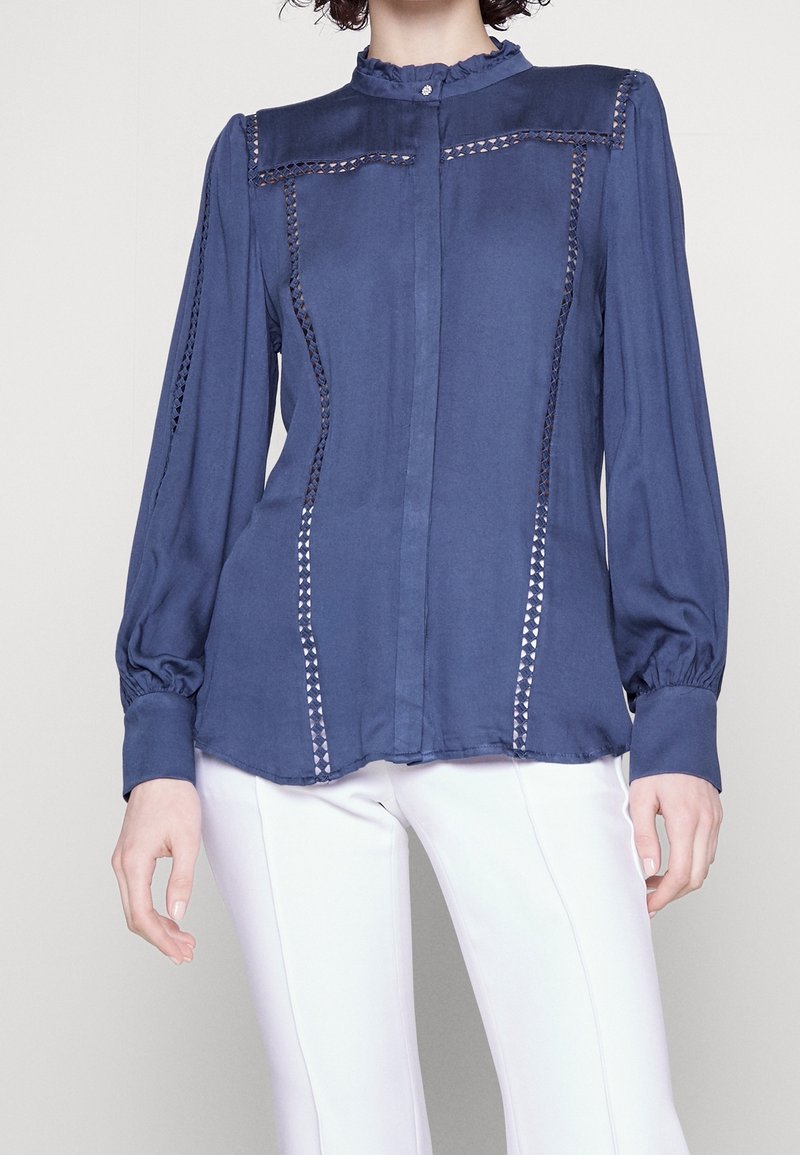 Femme portant un chemisier bleu à manches longues avec des découpes en dentelle et un pantalon blanc, montrée du cou aux hanches sur un fond uni.