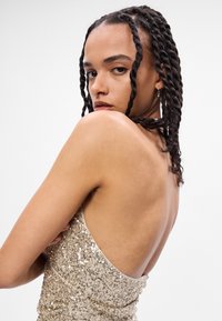 Χρυσό φόρεμα με παγιέτες, με χαμηλή πλάτη, λεπτές τιράντες και στενή γραμμή. Το ύφασμα έχει λεία υφή και λαμπερή φινέτσα.