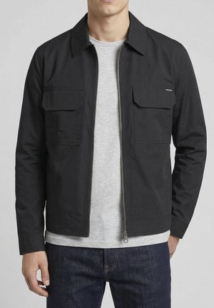 Man draagt een zwarte jas met rits en borstflapzakken over een lichtgrijs T-shirt en donkerblauwe spijkerbroek, staand tegen een effen achtergrond.