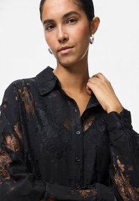 Young woman with dark hair wearing silver hoop earrings and a sheer black lace button-up shirt, holding her collar with one hand.
