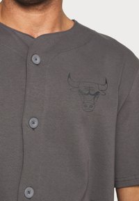 Šedé tričko s krátkým rukávem a zapínáním na knoflíky, na levé straně hrudi s černým logem býka a hladkou bavlněnou texturou.