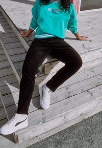 Persona che indossa una felpa turchese, pantaloni neri e sneakers bianche, seduta su gradini di legno all'aperto con le mani appoggiate dietro di sé.