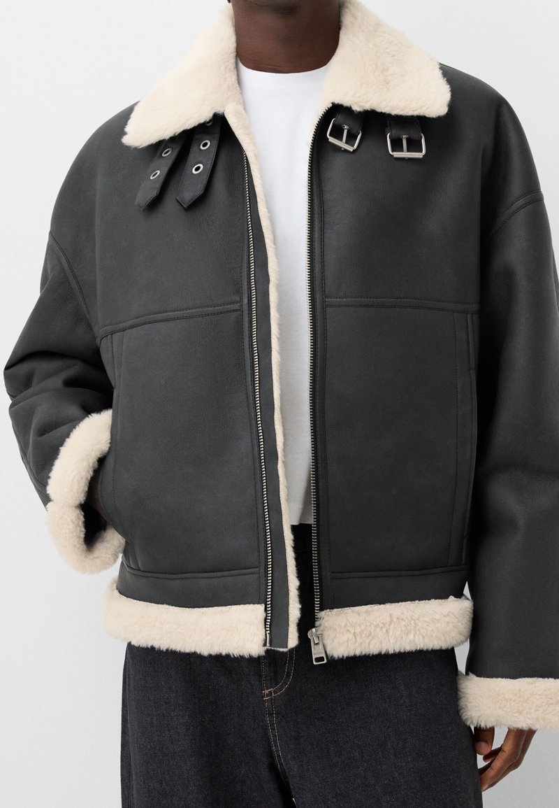 Veste en shearling noire avec col, poignets et ourlet en fourrure blanche, portée ouverte sur une chemise blanche et un jean foncé, main dans la poche.