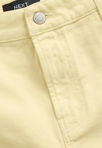 Tissu en denim jaune clair avec des nervures verticales, comportant un bouton argenté gravé "NEXT DENIM", avec des coutures nettes sur les bords.