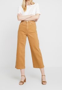 Pantalones de pierna ancha de tiro alto, en color beis, recortados, confeccionados en resistente algodón denim, con cierre de botón en la parte frontal y bolsillos laterales, combinados con una blusa blanca.