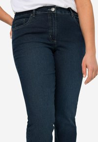 Tamsiai mėlyni džinsai su siauru siluetu, turintys vieną mygtuką uždarymui, priekinėmis kišenėmis ir kontrastinėmis siuvimo detalėmis.