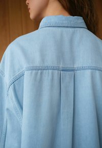 Lichtblauw denim overhemd met een klassieke kraag, plooidetail aan de achterkant en een soepele textuur. Eenvoudig ontwerp met zichtbare stiksels.