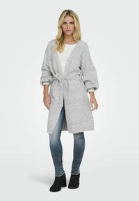 Cardigan en tricot gris clair avec une ceinture à nouer, épaules tombantes et manches retroussées, associé à un jean bleu et des bottines noires.