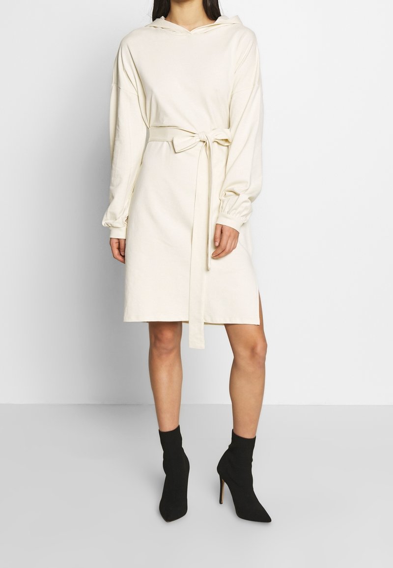 Vestido sudadera color crema con mangas largas y cinturón en la cintura. Presenta un ajuste relajado, aberturas laterales y una tela suave y texturizada.