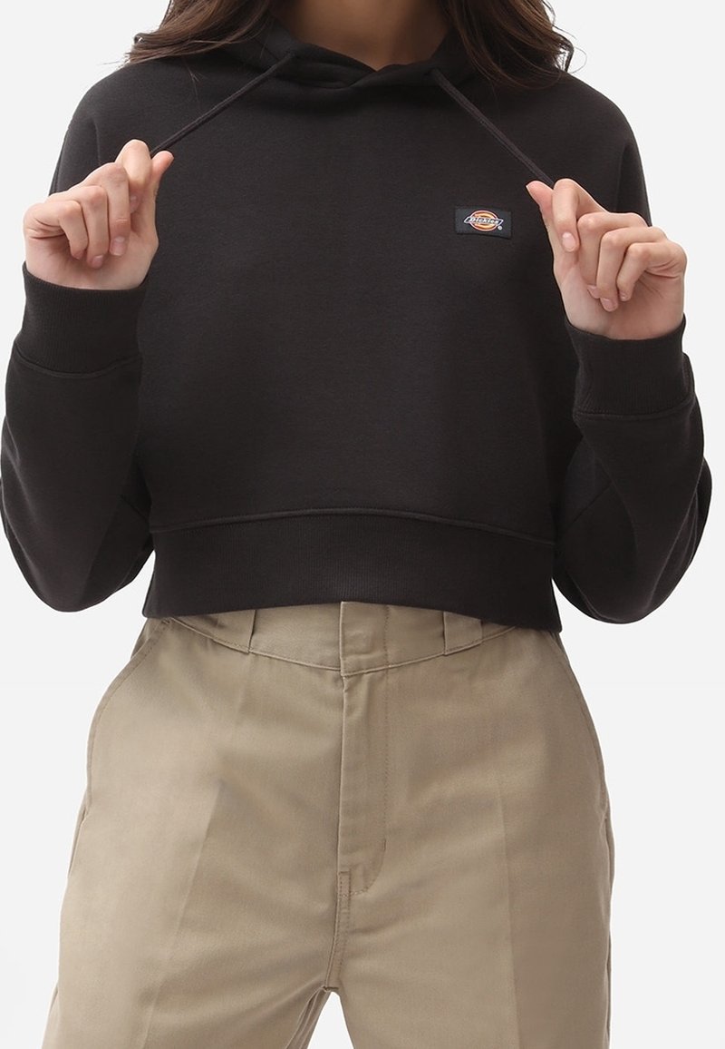 Sweat à capuche noir coupé avec un cordon de serrage, orné d'un petit patch logo. Associé à un pantalon beige taille haute, texture lisse visible.