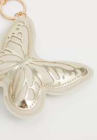 Portachiavi a forma di farfalla realizzato in pelle beige con dettagli in metallo dorato. Presenta motivi a rilievo sulle ali e un attacco con anello in metallo.