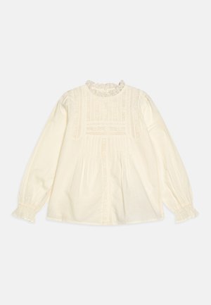 Blusa de cor creme com mangas longas, detalhes em renda, preguinhas e decote franzido. Apresenta um corte descontraído e uma textura de tecido suave.