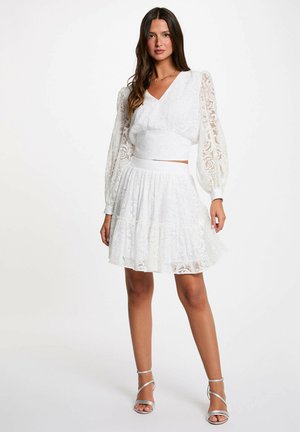 Donna che indossa un vestito bianco in pizzo a due pezzi con maniche lunghe a sbuffo e gonna svasata, abbinato a sandali argento con tacco alto.