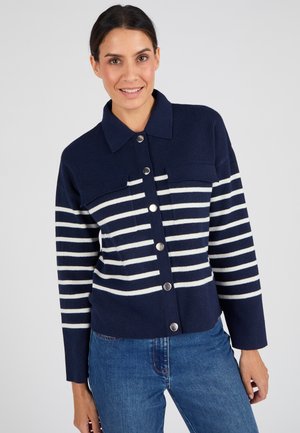 Femme souriante portant un cardigan bleu marine à rayures horizontales blanches et boutons argentés, associé à un jean bleu, sur un fond uni.