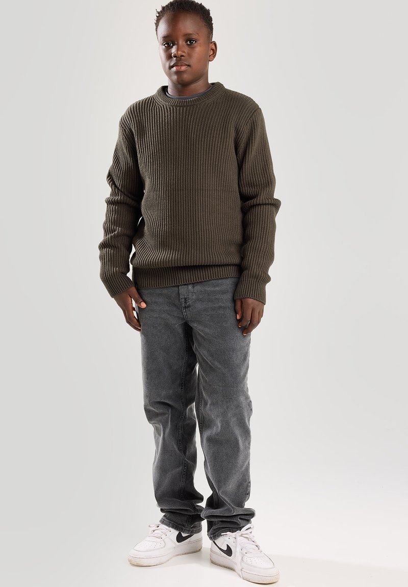 Olivenfarbener gerippter Pullover mit Rundhalsausschnitt, kombiniert mit ausgebleichten grauen Jeans und weißen Sneakers. Strukturierter Stoff, lässige Passform und entspannter Stil.