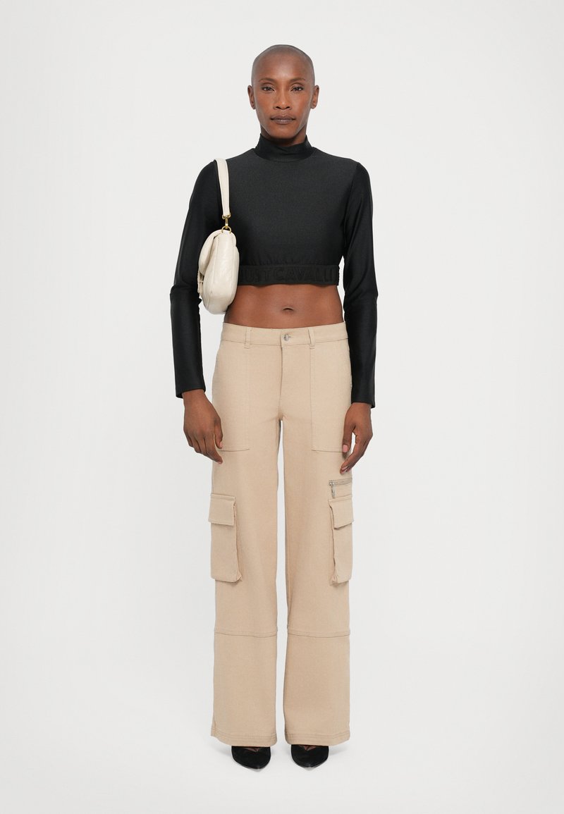 Black cropped long-sleeve top with textured band; beige cargo pants with side pockets and zipper; model holds a small cream handbag.