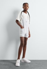 Stripete overallshorts i lyseblått og hvitt, med justerbare stropper, frontlomme og sidelommer, kombinert med en kremfarget t-skjorte og hvite joggesko.