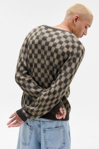 Pulover tricotat în carouri maro și negru, cu o croială lejeră, mâneci lungi și tivuri cu nervuri. Modelul poartă blugi de culoare albastru deschis.