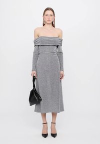 Rochie tricotată gri, cu umerii la vedere, cu mâneci lungi, textură cu nervuri și tăietură de lungime medie; asortată cu o geantă neagră și pantofi cu toc înalt.