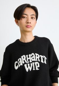 Czarny sweter z dzianiny z puszystym białym logo graficznym "Carhartt WIP" na wysokości klatki piersiowej, okrągłym dekoltem i luźnym krojem.