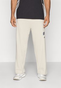 Homem vestido com calças de fato Adidas beje, sapatilhas brancas e uma camisa preta, contra um fundo claro e liso.