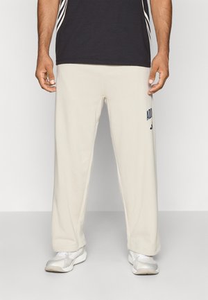 Bărbat îmbrăcat în pantaloni de trening bej Adidas, adidași albi și un tricou negru, pe un fundal simplu de culoare deschisă.