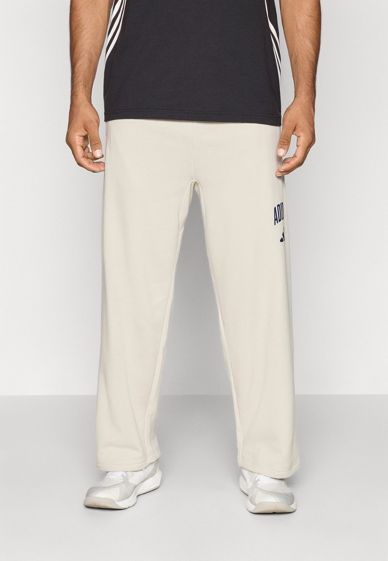 Homem vestido com calças de fato Adidas beje, sapatilhas brancas e uma camisa preta, contra um fundo claro e liso.