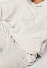 Persoon in een lichtbeige fleece hoodie en bijpassende broek, zittend met één arm gebogen en rustend op de knie, met een zachte textuur en naden zichtbaar.