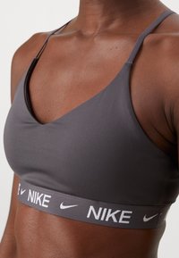 Sivi sportski brushalter s v-izrezom, tankim trakama i kontrastnim bijelim Nike logom duž elastičnog pojasa. Glatka tekstura.
