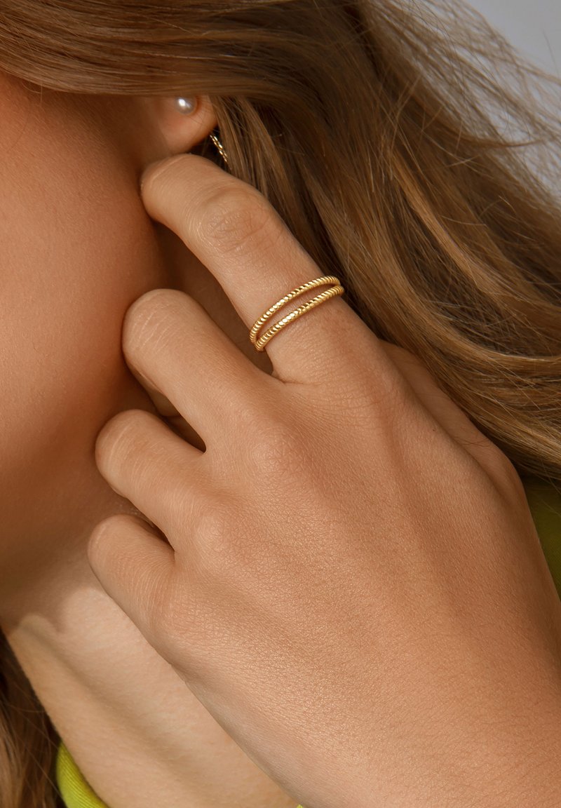 Goudkleurige ringen met gedraaide ontwerpen gedragen aan een hand. Een parel oorbel is zichtbaar in het oor, met gestructureerde huid in de buurt.