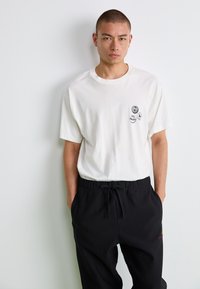 Tricou alb din bumbac cu guler rotund, având grafica imprimată neagră pe piept, asociat cu pantaloni negri cu șnur.