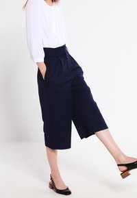 Navy blue cropped trousers with a high waist, pleated front, and side pockets, paired with a white blouse and low-heeled black shoes.