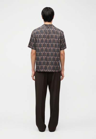 Άνδρας που στέκεται με την πλάτη γυρισμένη φορώντας κοντομάνικο πουκάμισο με σχέδιο και φαρδιά σκούρα καφέ παντελόνια σε απλό φόντο.