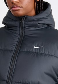 Mustmust mustpusa, millel on kõrge krae, esimesel külje lukul ja rinnal väike valge Nike logo. Sile ja läikiv tekstuur.