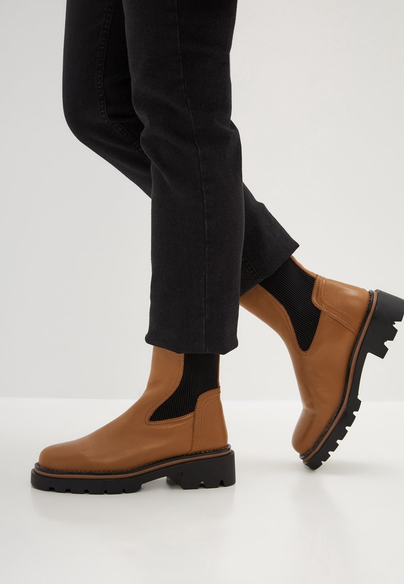 Bottines en cuir marron avec des panneaux latéraux élastiques noirs portées avec un pantalon noir, un pied légèrement levé montrant la semelle.
