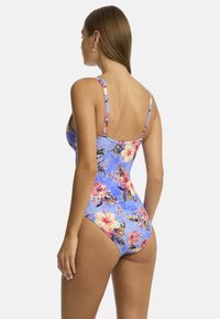 Een vrouw met lang haar kijkt van zich af, terwijl ze een blauwe bloemenbadkleding draagt versierd met roze en gele bloemen, wat een tropisch ontwerp laat zien.